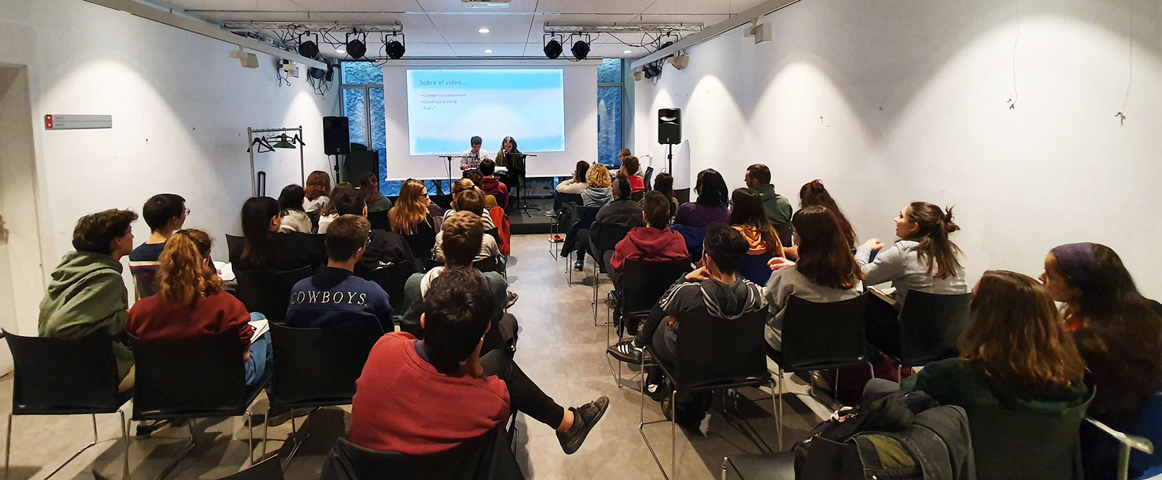 Fotografia general de les jornades on veiem tot l'espai ple de cadires amb les assistents i al fons, a l'escenari, dues persones amb un micròfon fent la xerrada inagural.